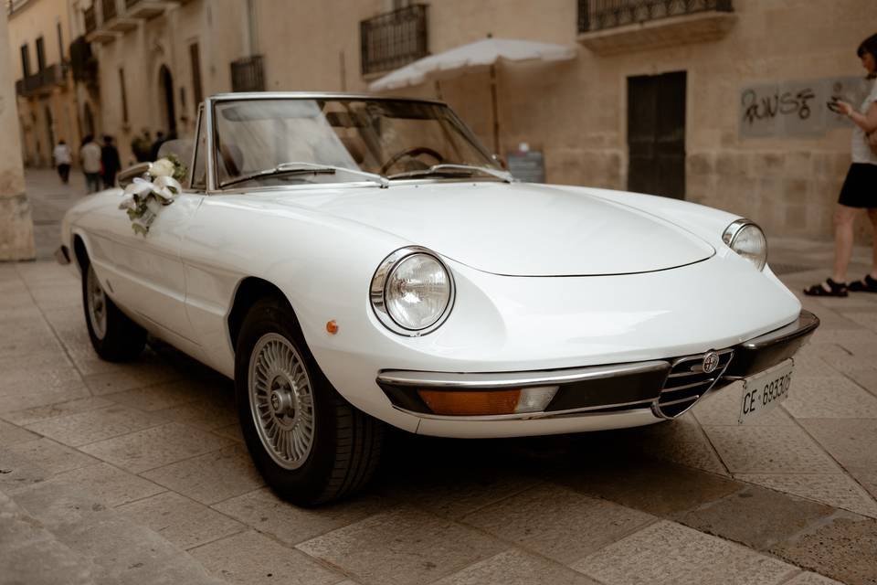 L'Alfa Romeo Spider (Duetto) bianca, cabrio, con un mazzo di fiori sulla portiera, in una strada lastricata